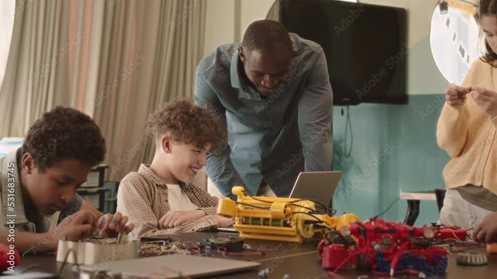 Medium slowmo of group of diverse schoolkids constructing robots on desk in classroom, working on electronics project together Medium slowmo of group of diverse schoolkids constructing robots on desk in classroom, working on electronics project together