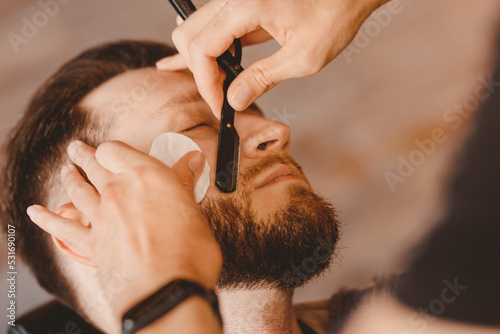 Hipster man sitting in armchair barber shop hairdresser shaves his beard with dangerous razor, vintage color