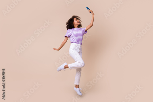 Full length photo of sweet cute girl dressed purple t-shirt jumping high recording video vlog modern gadget isolated beige color background