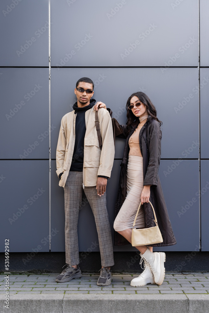 Leinwandbild Motiv - LIGHTFIELD STUDIOS : full length of stylish interracial couple in autumnal outfits and trendy sunglasses standing near building.