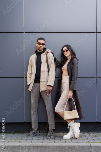 Photographie full length of stylish interracial couple in autumnal outfits and trendy sunglasses standing near building