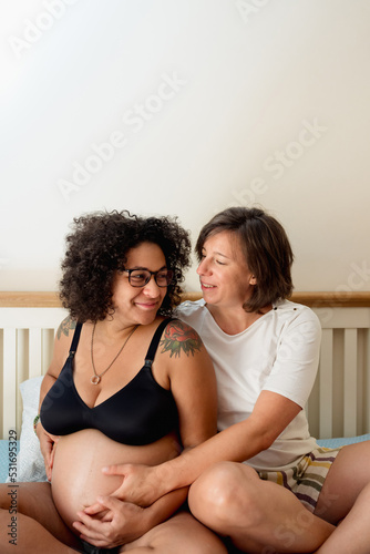 Woman embracing her pregnant girlfriend while sitting on the bed together at home. Pregnancy and lgbt concept.