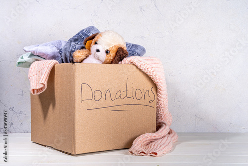 Donation box with clothes, toys and food on white table background. Cardboard donation box full with toys, female and child clothes, foods. Social care, volunteering, charity concept copy space photo