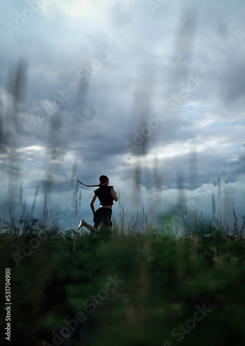 Woman with ponytail in black skirt and shirt runs barefeet through a field with tall grass under a dark cloudy sky. 3D render.