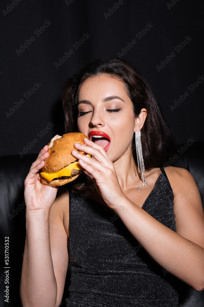 stylish and elegant woman with closed eyes eating burger isolated on black.