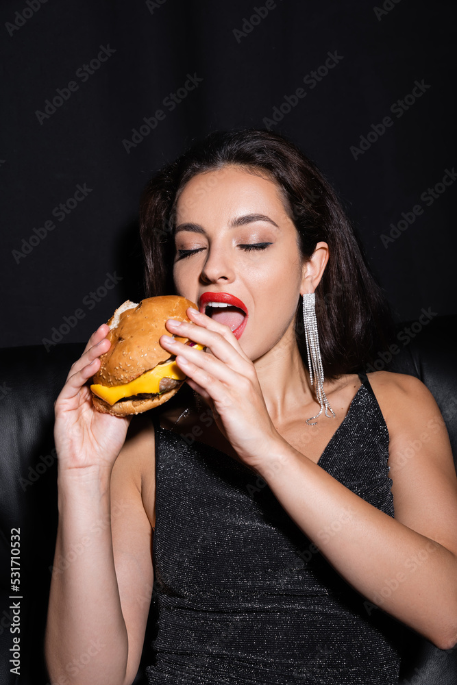 stylish and elegant woman with closed eyes eating burger isolated on black. stylish and elegant woman with closed eyes eating burger isolated on black.