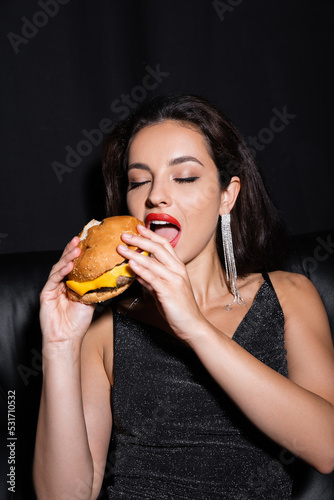 stylish and elegant woman with closed eyes eating burger isolated on black.