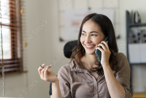 Happy asian businesswoman sitting at desk talking with mobile phone with somebody in office workplace.