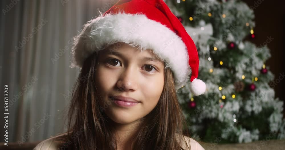 Portrait of a cute child in festive Christmas, portrait little girl in a Christmas hat Santa Claus sitting on sofa at home looking at camera on the background decorative Christmas tree. Portrait of a cute child in festive Christmas, portrait little girl in a Christmas hat Santa Claus sitting on sofa at home looking at camera on the background decorative Christmas tree.