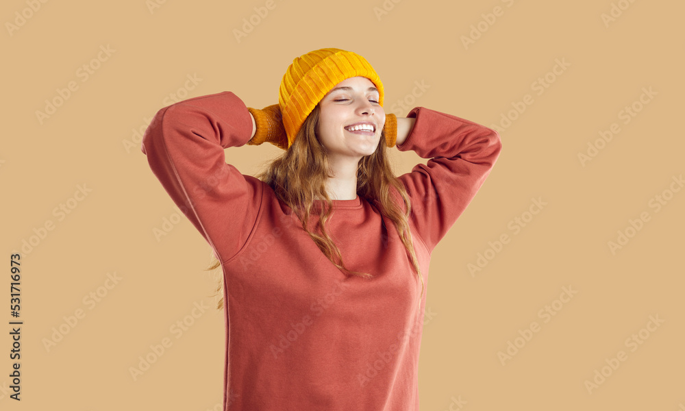 Happy relaxed attractive young woman in cozy yellow hat, gloves and trendy dark coral sweatshirt puts hands behind head, closes eyes, breathes fresh air and smiles. Studio shot. Autumn fashion concept Happy relaxed attractive young woman in cozy yellow hat, gloves and trendy dark coral sweatshirt puts hands behind head, closes eyes, breathes fresh air and smiles. Studio shot. Autumn fashion concept