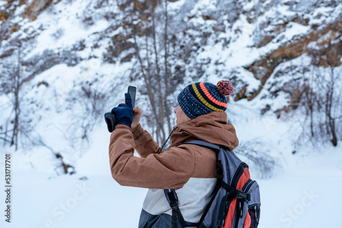 young man in brown warm clothes with backpack walking in snow among rocks and cliffs in winter looking at view taking photos with smartphone Active lifestyle hiking