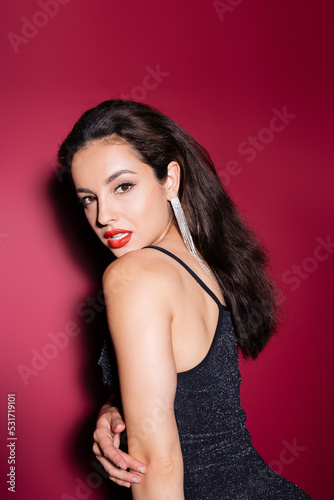 brunette woman in shiny earring and black lurex dress looking at camera on red background.