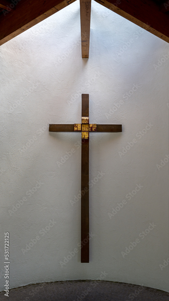 Braunes Holzkreuz mit Golddekor an einer weißen halbrunden Wand, darüber eine Holzdecke mit Lichteinlass