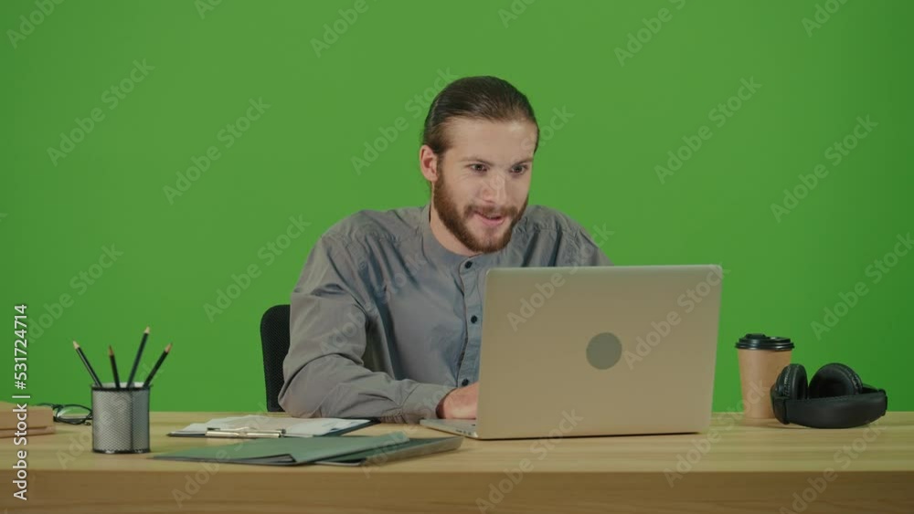 Young Motivated Bearded Student Typing on Laptop Working in Internet,Doing Online Job and Browsing Site at Work Table,Smiling and Making Winning Gesture on Green Screen,Chroma Key.Remote Work Concept Young Motivated Bearded Student Typing on Laptop Working in Internet,Doing Online Job and Browsing Site at Work Table,Smiling and Making Winning Gesture on Green Screen,Chroma Key.Remote Work Concept