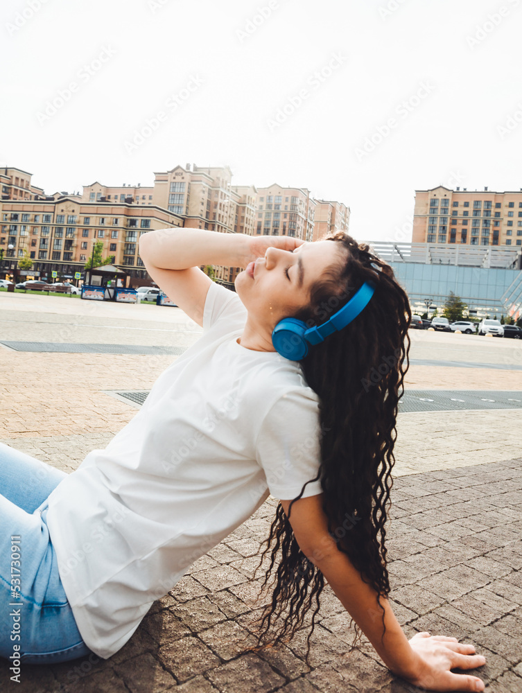 A smiling girl with dreadlocks in headphones is sitting on the floor in the park. A happy young woman relaxing with headphones, enjoying music. Space for copying.generation z