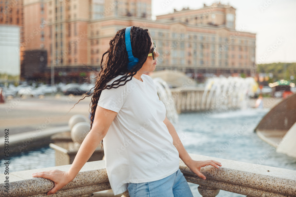 A young cheerful happy woman with dreadlocks, dressed in a white T-shirt, dancing, listening to music with headphones, resting, relaxing in a city park, walking along an alley. Urban lifestyle concept A young cheerful happy woman with dreadlocks, dressed in a white T-shirt, dancing, listening to music with headphones, resting, relaxing in a city park, walking along an alley. Urban lifestyle concept