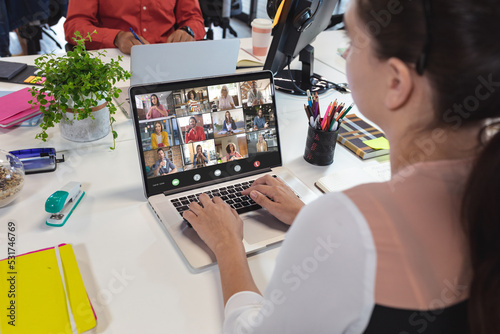 Rear view of caucasian woman video conferencing with coworkers over laptop at office