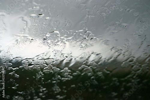 Glasscheibe mit Regentropfenmuster vor grüner Landschaft und diesigem grau-weißen Himmel bei Regen am Morgen im Herbst