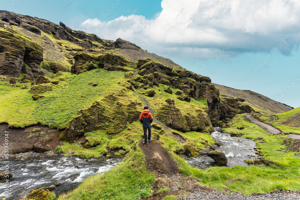 Tourist man standing on Kvernufoss waterfall with mossy rocky canyon in summer on sunny day at Iceland