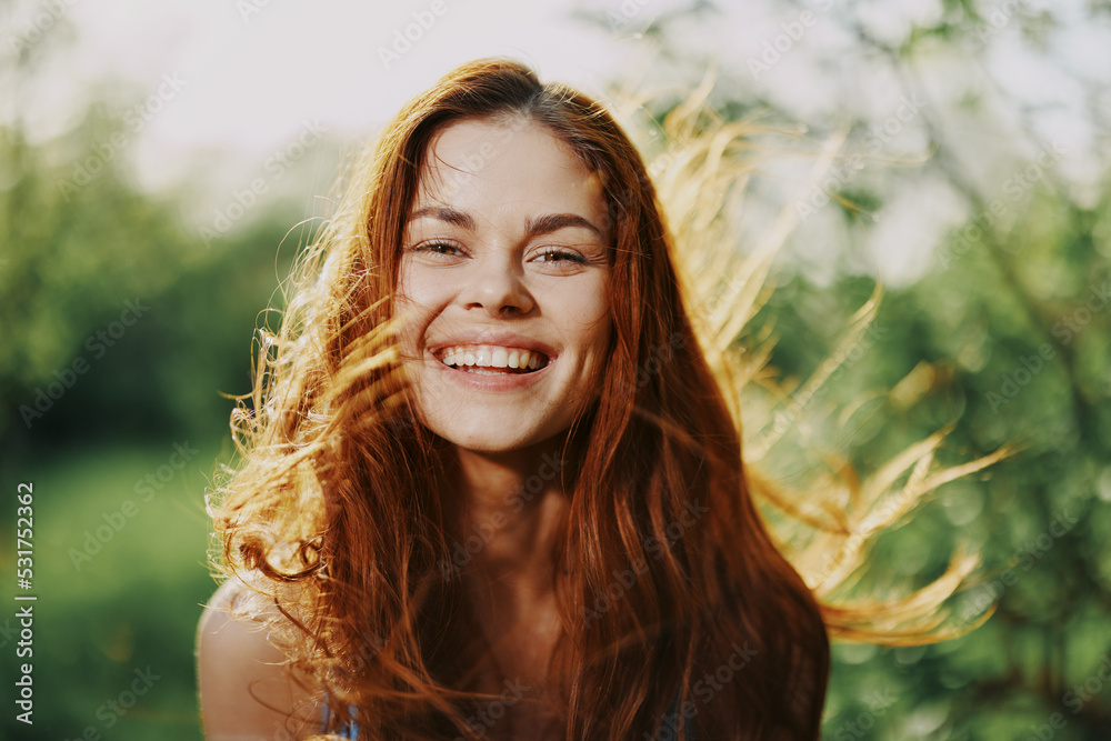 Naklejka premium Woman portrait smile happiness catch looks into the camera with a smile with teeth spring flying hair long red, the concept of health and beauty hair sunset