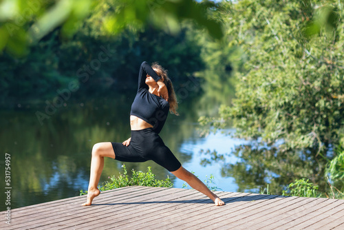 A woman in sports black clothes practicing yoga performs a variation of the virabhadrasana exercise, warrior pose, trains on a warm summer morning while standing on a wooden bridge near the river