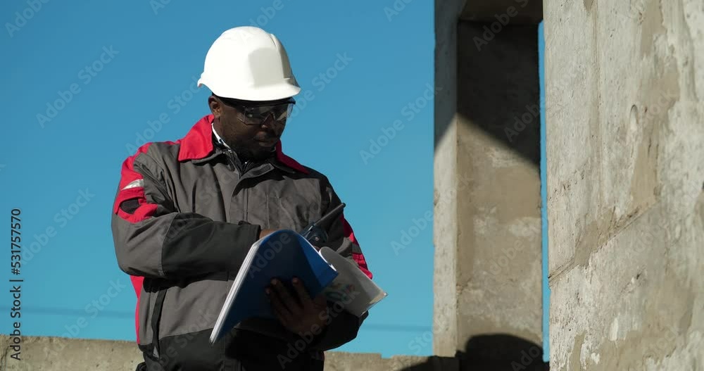 African american work superintendent stands at construction site, looks in work papers and speaks via portable radio transmitter. Cinema 4K 60fps video African american work superintendent stands at construction site, looks in work papers and speaks via portable radio transmitter. Cinema 4K 60fps video
