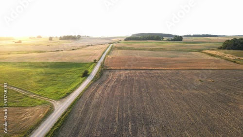 Agriculture and nature concept. Aerial landscape view of various crop fields and rural road during late summer sunset. Image contains noise and blurriness. In horizon visible electric windmills