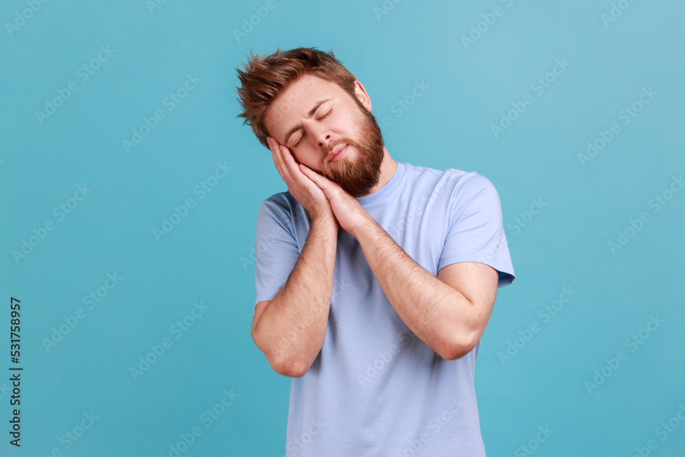 Portrait of tired bearded man sleeping laying down on his palms and smiling pleased, having comfortable nap and resting, dozing off. Indoor studio shot isolated on blue background. Portrait of tired bearded man sleeping laying down on his palms and smiling pleased, having comfortable nap and resting, dozing off. Indoor studio shot isolated on blue background.