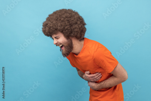 Φωτογραφία Side view of laughing man with Afro hairstyle in orange T-shirt holding his stomach and hunched in crazy hysterical laughter, sincere joyful emotions