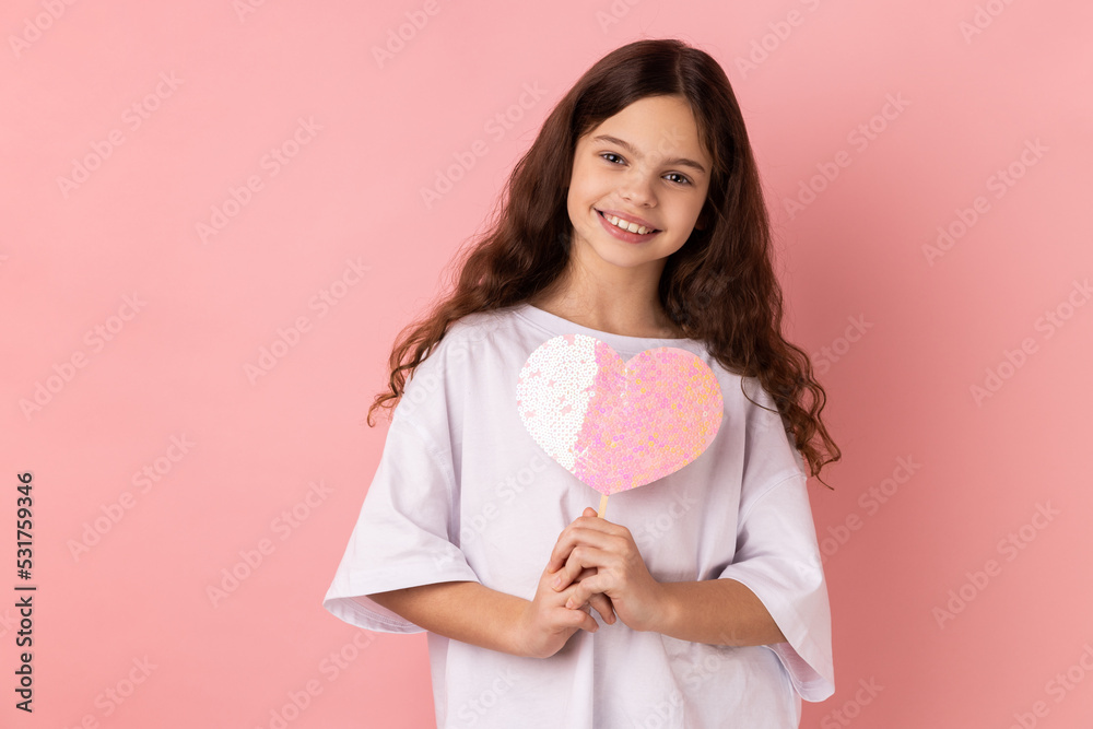 Portrait of happy satisfied little girl wearing white T-shirt holding paper pink heart on stick and smiling to camera, sharing love. Indoor studio shot isolated on pink background. Portrait of happy satisfied little girl wearing white T-shirt holding paper pink heart on stick and smiling to camera, sharing love. Indoor studio shot isolated on pink background.