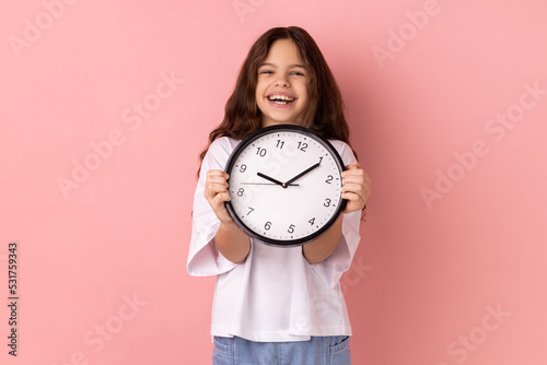 Portrait of smiling delighted little girl wearing white T-shirt holding wall clock, being happy, deadline, satisfied with completed home task. Indoor studio shot isolated on pink background.