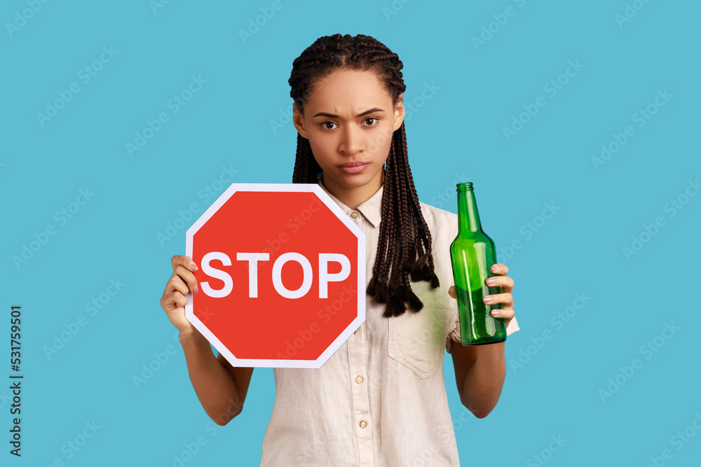 Portrait of serious woman with dreadlocks holding red stop sign and bottle with alcoholic beverage, calls on dont drink alcohol, wearing white shirt. Indoor studio shot isolated on blue background. Portrait of serious woman with dreadlocks holding red stop sign and bottle with alcoholic beverage, calls on dont drink alcohol, wearing white shirt. Indoor studio shot isolated on blue background.