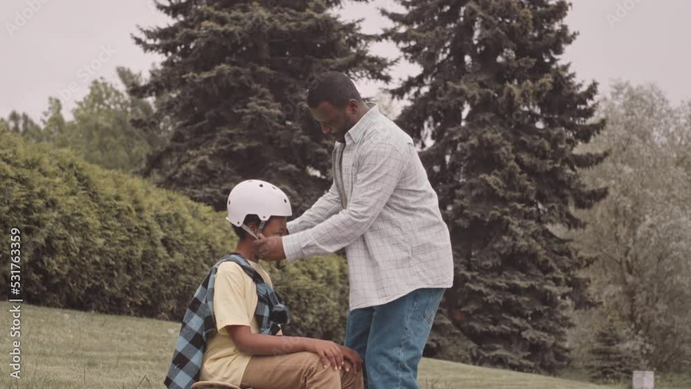 Slowmo of Black man putting on helmet on head of his teenage son while teaching him to ride skateboard outdoors in park Slowmo of Black man putting on helmet on head of his teenage son while teaching him to ride skateboard outdoors in park
