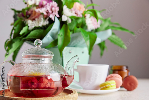 Natural herbal raspberry red tea in glass teapot, macaroons and bouquet of flowers. Aesthetic home breakfast. Self care, relaxing, wellness lifestyle.