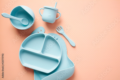 Baby feeding equipment. Flat lay silicone bib, plate, cup, cutlery on pastel beige background. Toddler self feeding training, baby weaning concept.