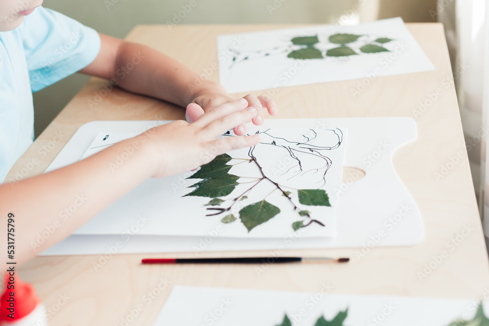 Cute child sitting at desk and making picture from dry birch leaves Cute child sitting at desk and making picture from dry birch leaves