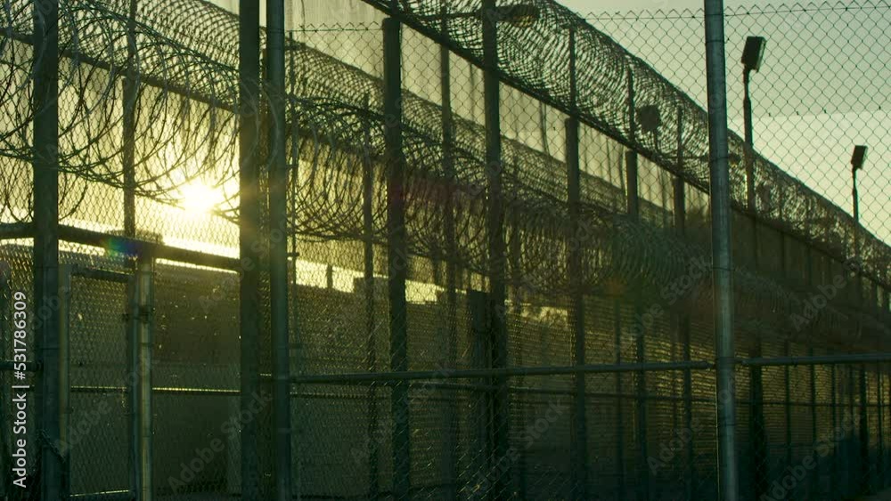 Overview of the electric chain-link fence with barbed wire of the prison, High barbed wire fence around prison or secret military base, detention facility, watchtower, last sun rays penetrating Overview of the electric chain-link fence with barbed wire of the prison, High barbed wire fence around prison or secret military base, detention facility, watchtower, last sun rays penetrating