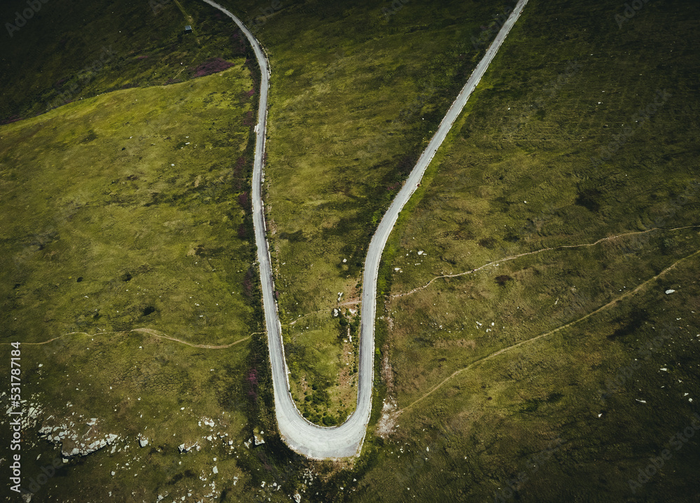 Aerial photo of mountain curved roadunder the mountain peak in  Europe. Alpine road - beautiful scenic landscape with green meadows and rocks from above. Dark and moody photo of a way in mountains.