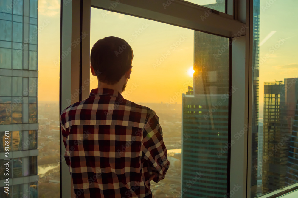 Back view of man in plaid shirt looking at cityscape through window of skyscraper. Summer time, sunset, warm illumination. Success, opportunity, sightseeing, discover and future concept Back view of man in plaid shirt looking at cityscape through window of skyscraper. Summer time, sunset, warm illumination. Success, opportunity, sightseeing, discover and future concept