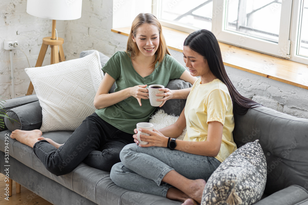 © ArtSys - Two millennial girl friends at home on the sofa talking, drinking coffee, discussing. Diverse friends spending fun time together and laughing, chat and talk. Asian and Caucasian model. © ArtSys - Two millennial girl friends at home on the sofa talking, drinking coffee, discussing. Diverse friends spending fun time together and laughing, chat and talk. Asian and Caucasian model.