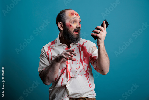 Spooky halloween monster holding smartphone in studio, using mobile phone app while being aggressive and scary. Frightening undead zombie with bloody wounds, cruel corpse with telephone. photo