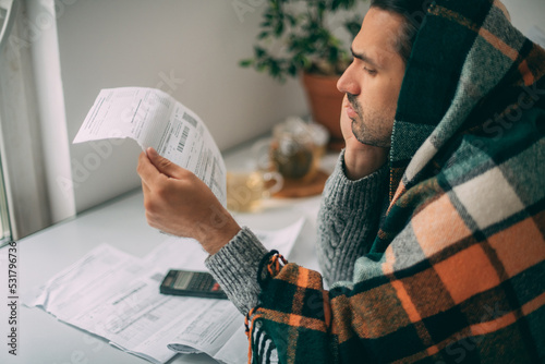 A sad man in a warm plaid is sitting at a table with utility bills in his hands. photo