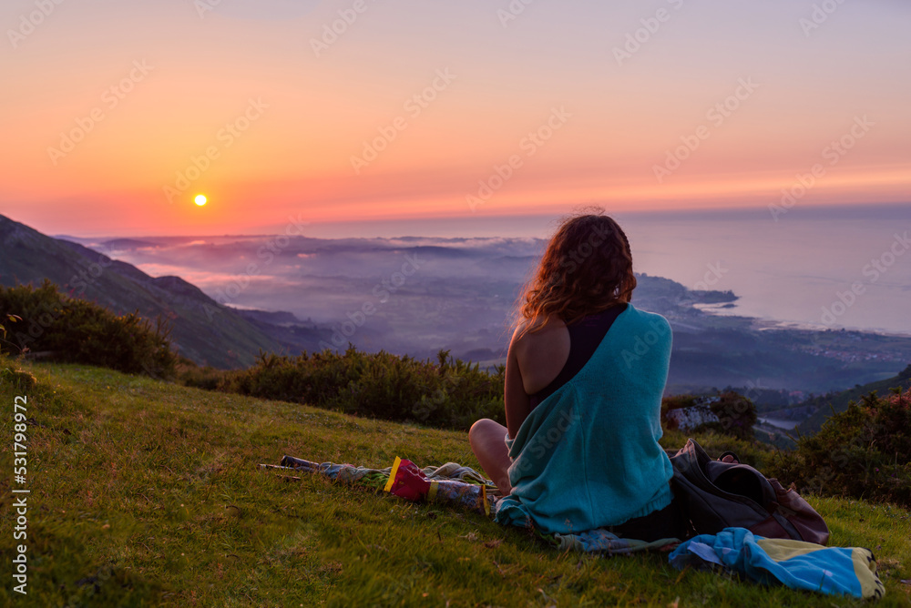 woman from behind, sitting contemplating the sunset from a mountain facing the sea.