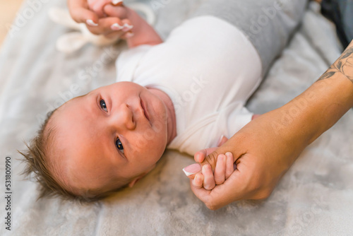 A couple months old baby boy with cute brown hair, laying on his back on a grey blanket, with his arms raised up, holding his mothers hands big fingers. High quality photo