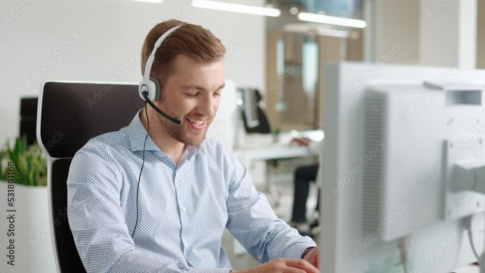 Close up of young handsome happy male worker sitting at desk in cabinet and typing on computer while talking in headset on call. Positive Caucasian man manager browsing on computer working in support