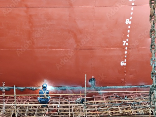 A red ship hull with draft mark and a man or worker who's sitting on the staging and welding on ship hull