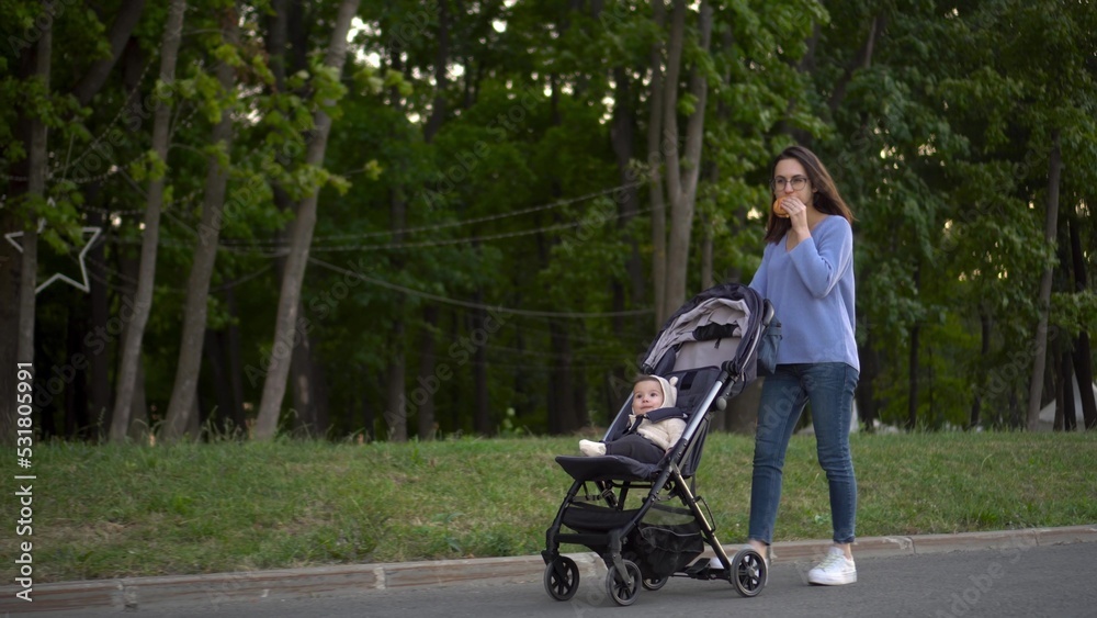 Mom walks with a baby in a stroller and eats a burger on the go. A young woman in glasses pushes a stroller with her son and eats a burger in the park.