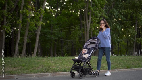 Mom walks with a baby in a stroller and eats a burger on the go. A young woman in glasses pushes a stroller with her son and eats a burger in the park.