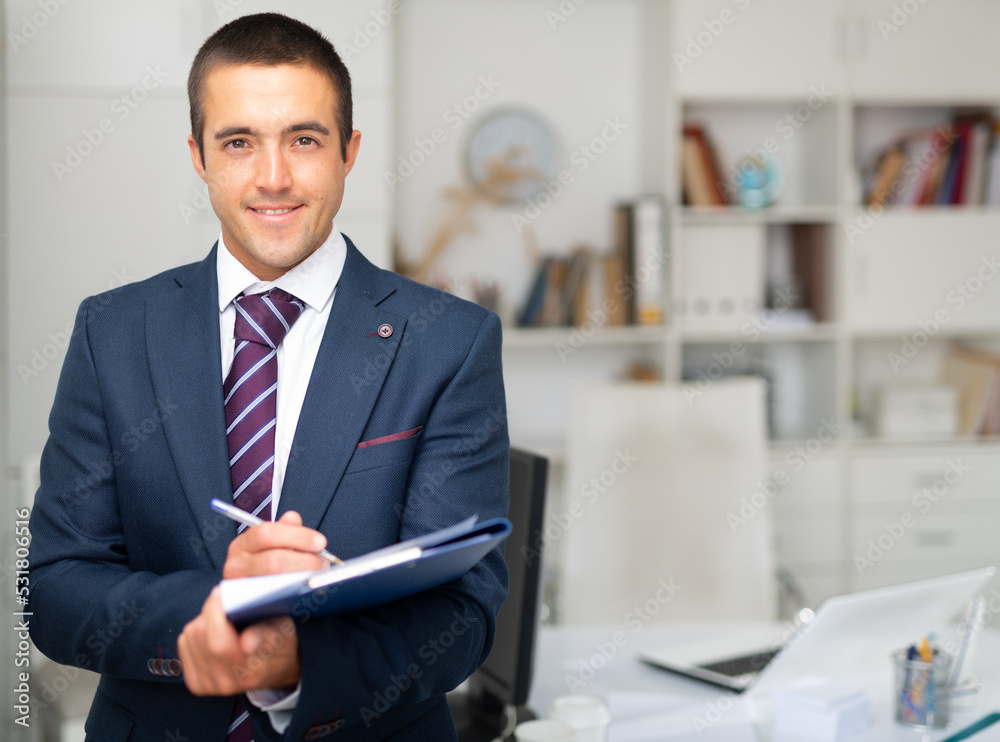 Portrait of confident smiling business assistant standing in office with clipboard, noting tasks