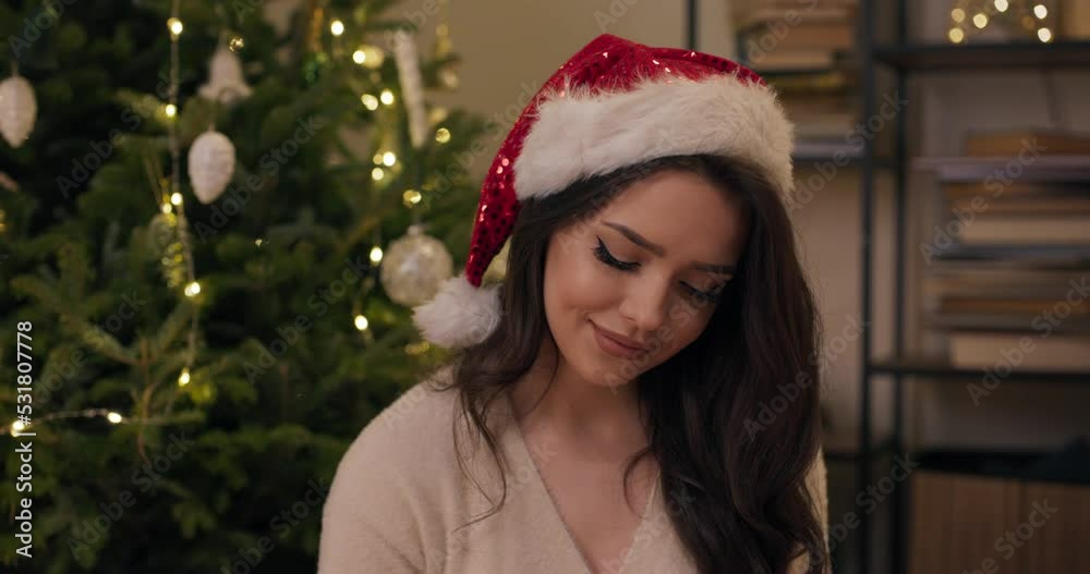 Winter evening. Close-up of an attractive brunette sitting by Christmas tree looking at prepared gifts. She is smiling, wearing beige sweater and Santa Claus hat. Winter evening. Close-up of an attractive brunette sitting by Christmas tree looking at prepared gifts. She is smiling, wearing beige sweater and Santa Claus hat.